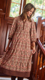 A woman wearing a brown and beige patterned tunic dress with three-quarter sleeves, standing on wooden stairs next to a wooden railing.