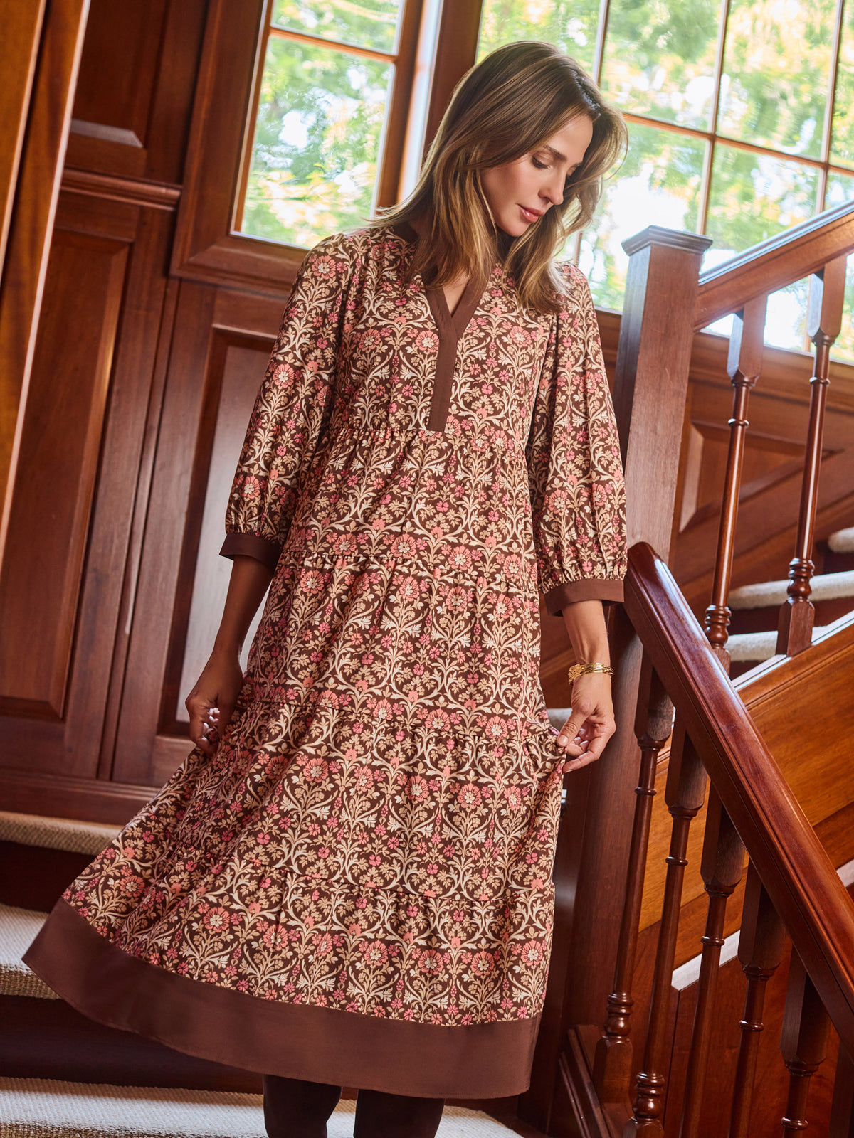 A woman wearing a brown and beige patterned tunic dress with three-quarter sleeves, standing on wooden stairs next to a wooden railing.
