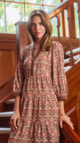 A woman wearing a brown and beige patterned tunic dress with three-quarter sleeves, standing on wooden stairs next to a wooden railing.