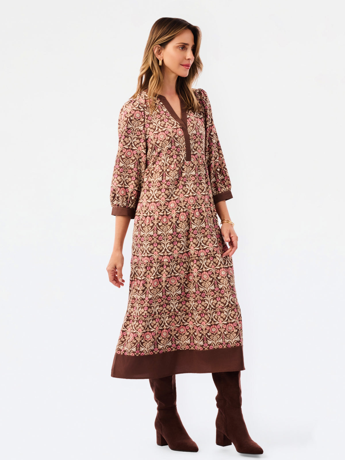 A woman modeling a long-sleeved, knee-length printed tunic dress in pink, brown, and beige, paired with matching brown boots, against a plain white studio background.