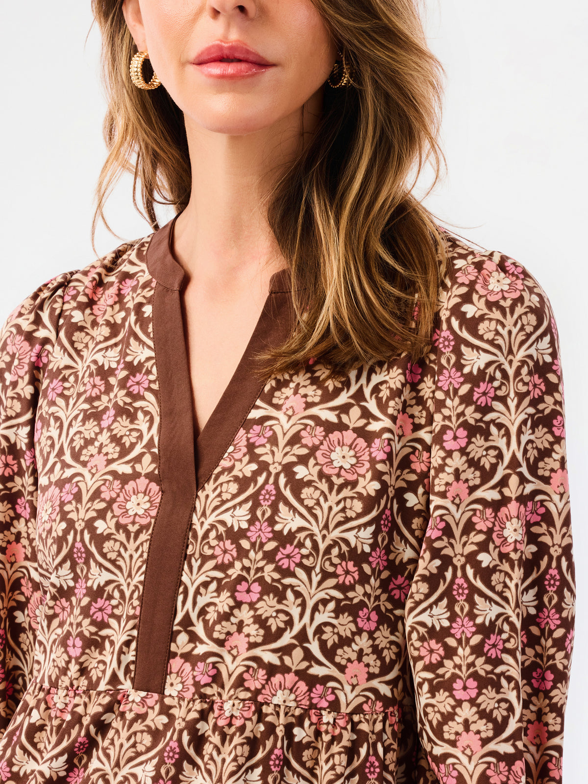 A close-up of a woman wearing a brown long-sleeve dress with a pink and beige floral pattern and a brown tie-neck detail.