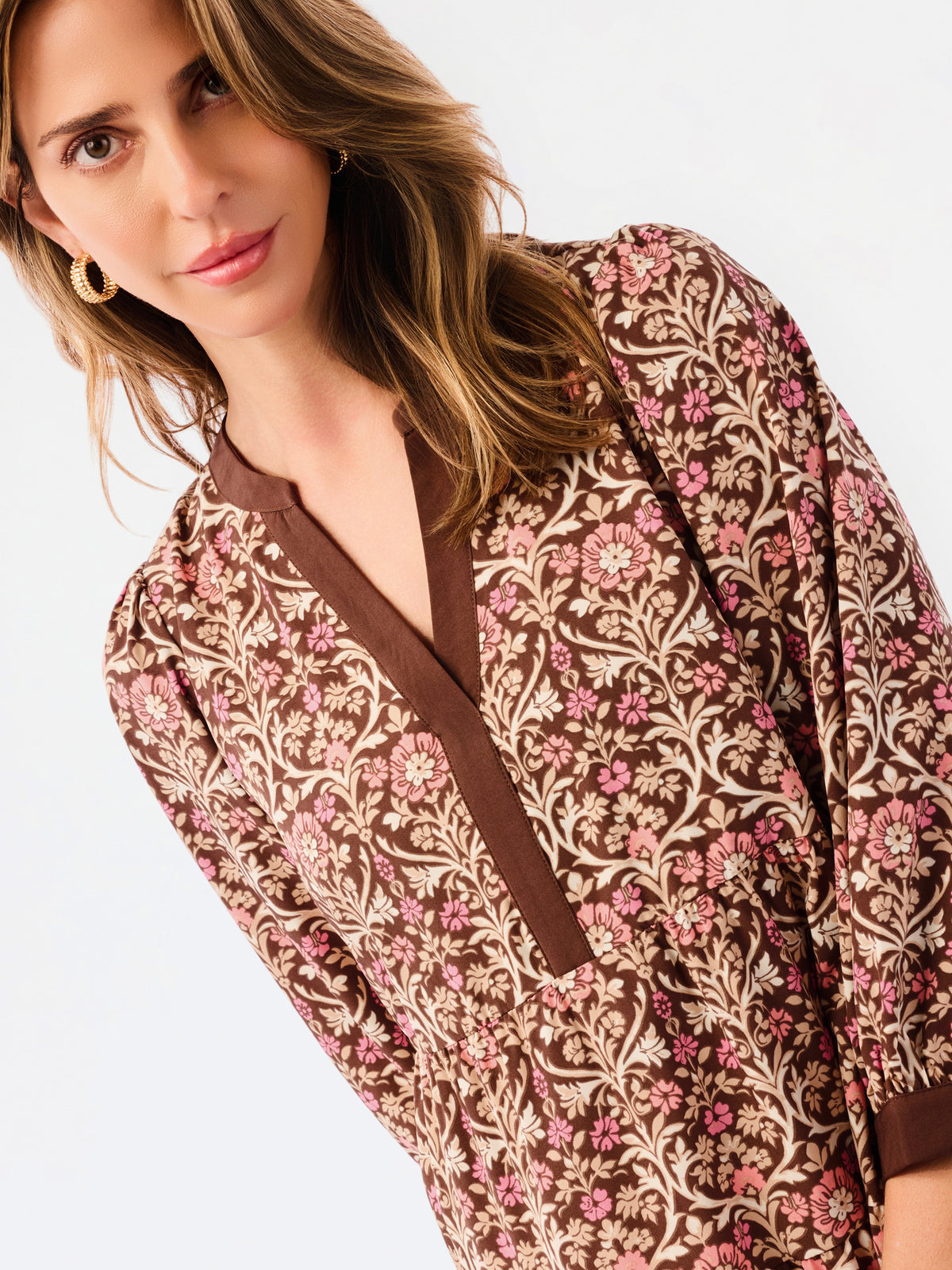A close-up of a woman wearing a brown long-sleeve dress with a pink and beige floral damask pattern and a brown V-neckline.