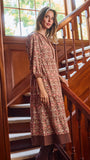 A woman wearing a brown and beige patterned tunic dress with three-quarter sleeves, standing on wooden stairs next to a wooden railing.