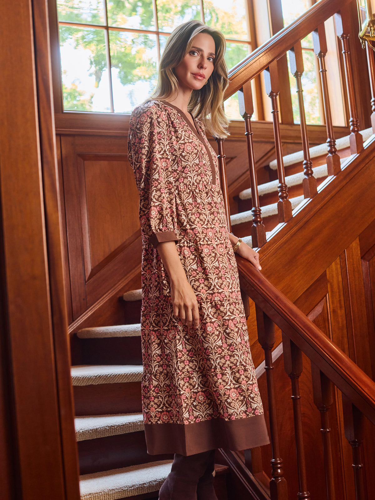 A woman wearing a brown and beige patterned tunic dress with three-quarter sleeves, standing on wooden stairs next to a wooden railing.