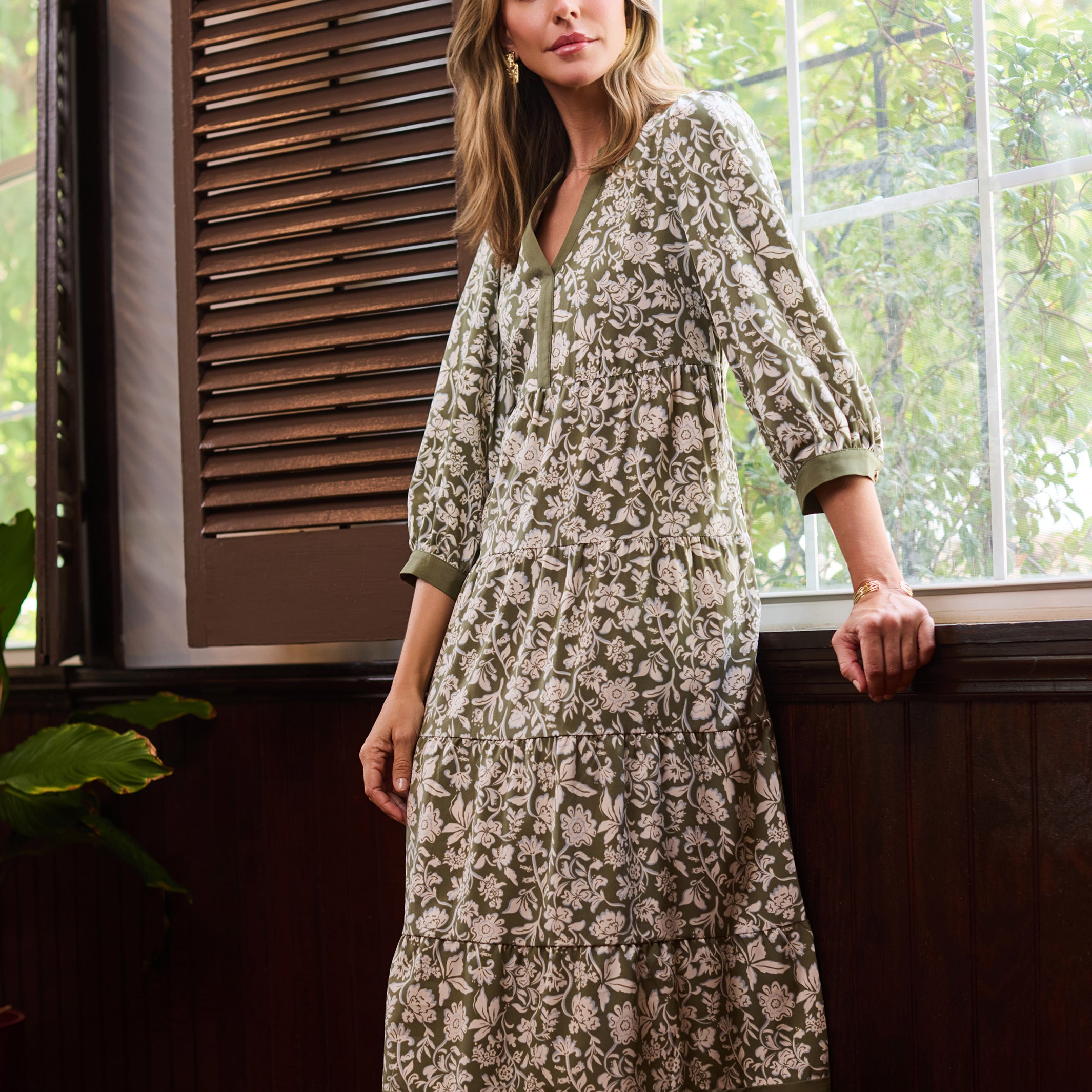 A woman models a long-sleeved, knee-length kurta with an all-over floral block print in beige and olive green, standing by a window in a rustic room.