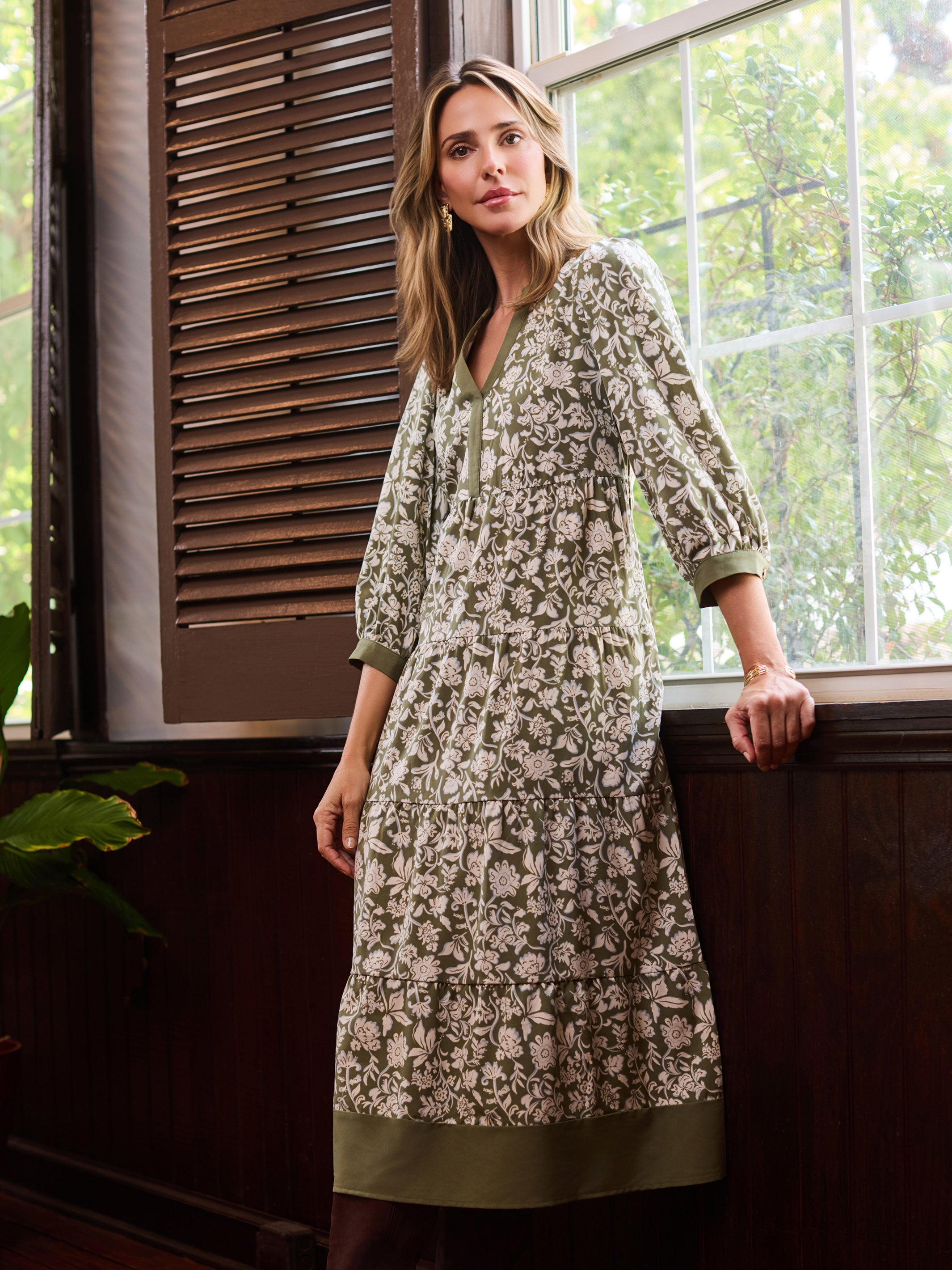 A woman models a long-sleeved, knee-length kurta with an all-over floral block print in beige and olive green, standing by a window in a rustic room.