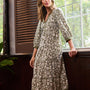 A woman models a long-sleeved, knee-length kurta with an all-over floral block print in beige and olive green, standing by a window in a rustic room.