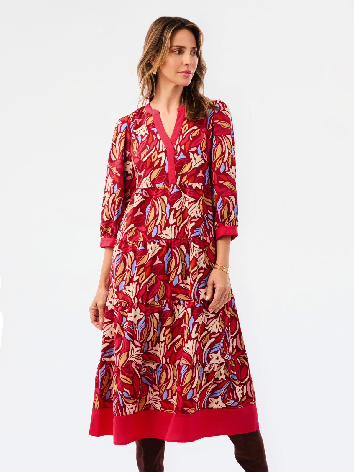 A woman modeling a red midi dress with a colorful abstract pattern in beige, blue, and gold, featuring a V-neckline and 3/4 sleeves, set against a plain white studio background.
