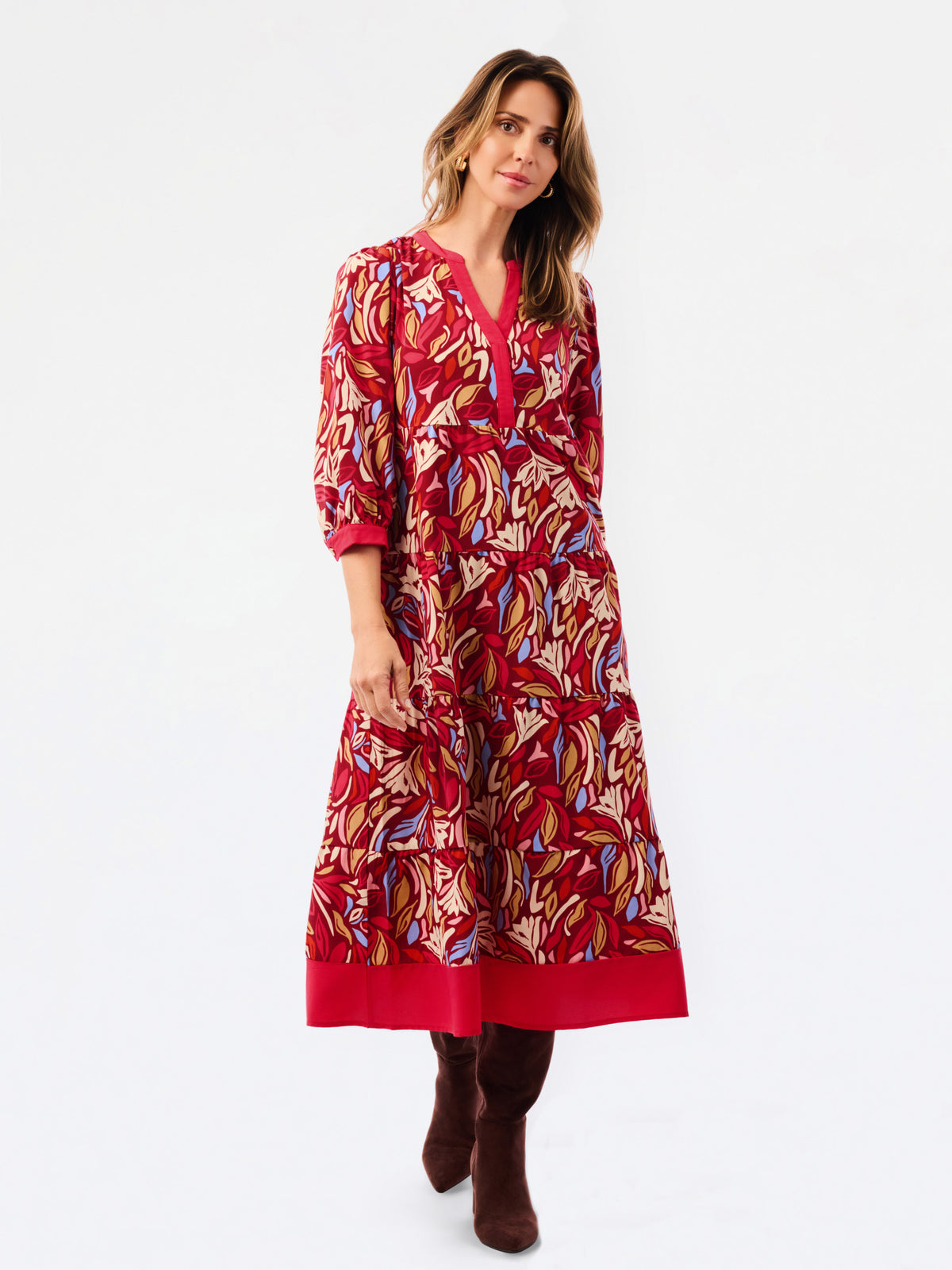 A woman modeling a red midi dress with a multicolor abstract print in beige, blue, and brown, featuring three-quarter sleeves and a V-neckline, paired with brown boots against a white studio background.