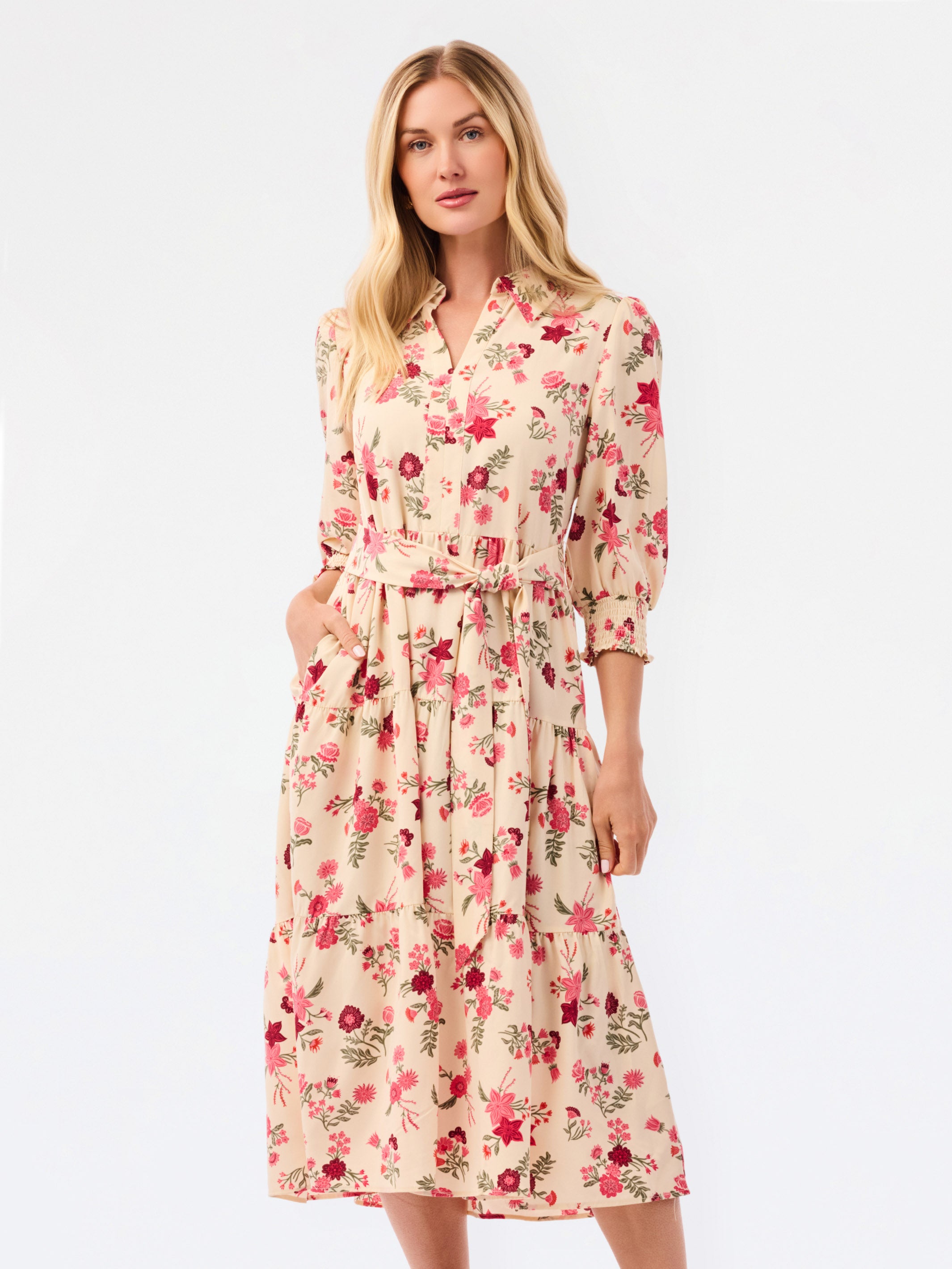 A woman modeling a beige midi shirt dress with a red and pink floral print, featuring a tie waist and 3/4 sleeves, set against a plain white studio background.