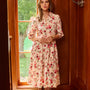 A woman stands in a warm wood-paneled room wearing a cream midi dress with a red and pink floral print, paired with brown boots.