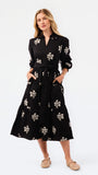 A woman modeling a black midi shirt dress with a white floral pattern, featuring a tie waist, long sleeves, and front pockets, paired with beige flat shoes against a white studio background.