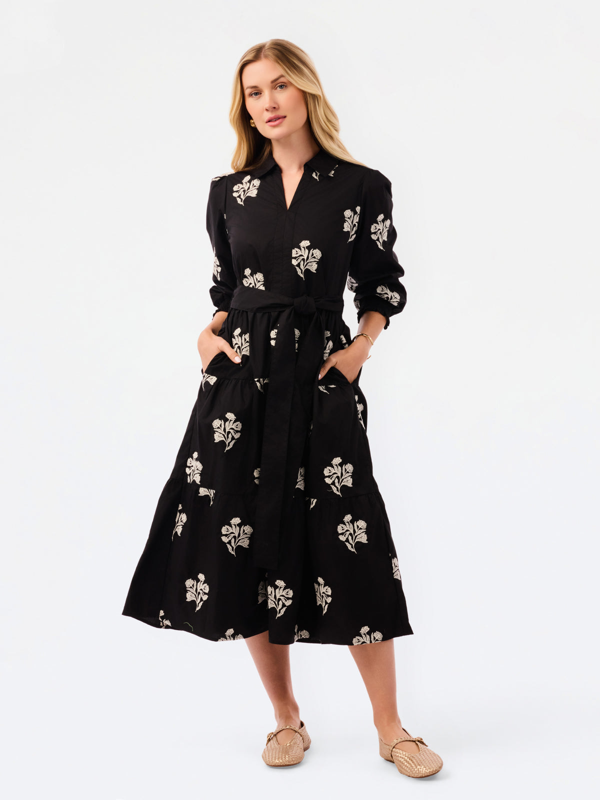 A woman modeling a black midi shirt dress with a white floral pattern, featuring a tie waist, long sleeves, and front pockets, paired with beige flat shoes against a white studio background.