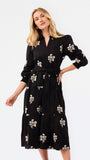 A blonde model wearing a black long-sleeve midi wrap dress with a white floral print, standing against a plain white studio background.
