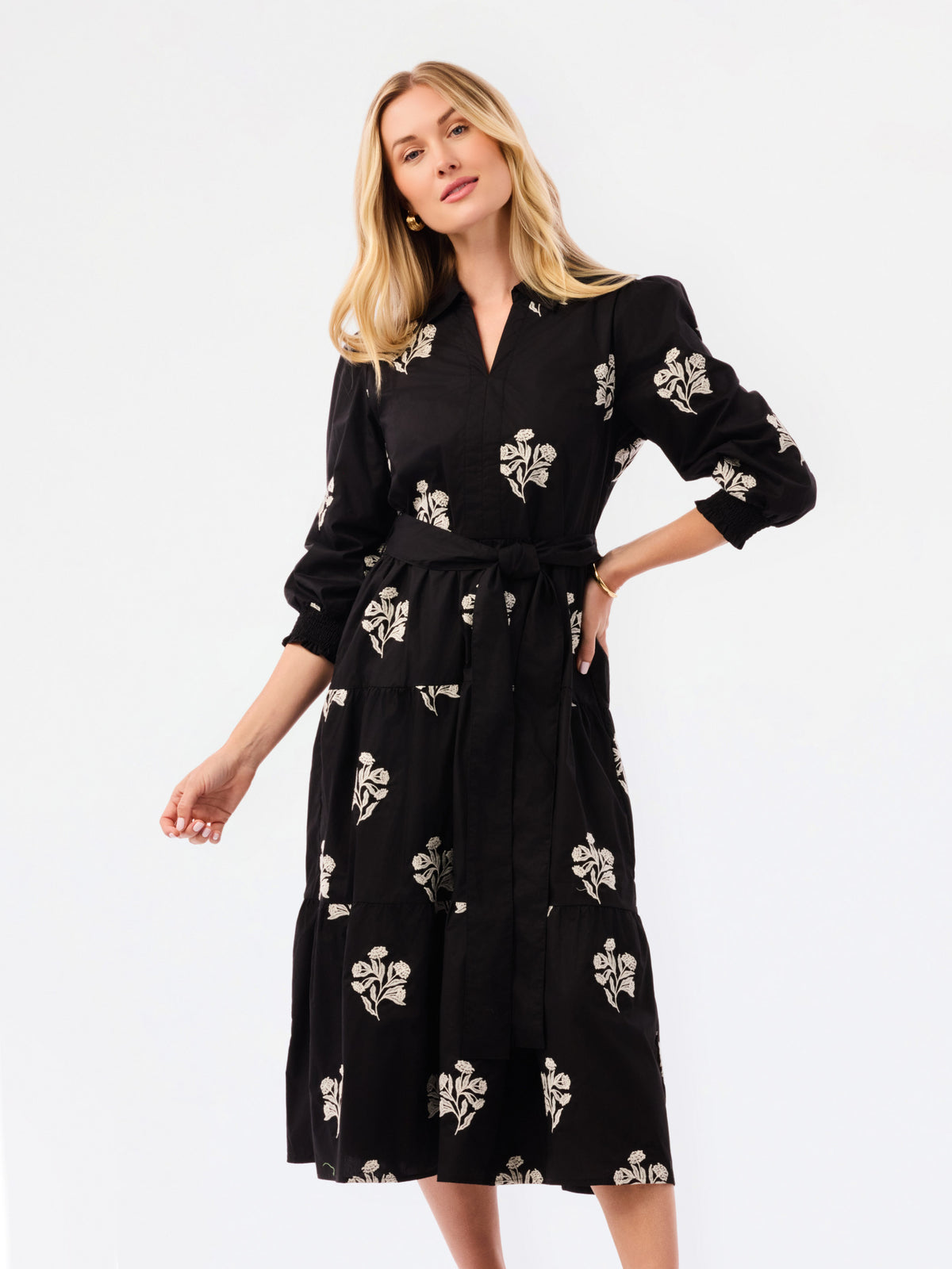 A blonde model wearing a black long-sleeve midi wrap dress with a white floral print, standing against a plain white studio background.