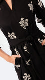 A close-up of a black long-sleeve dress with white floral embroidery, featuring a tie waist belt and side pockets, worn by a model.