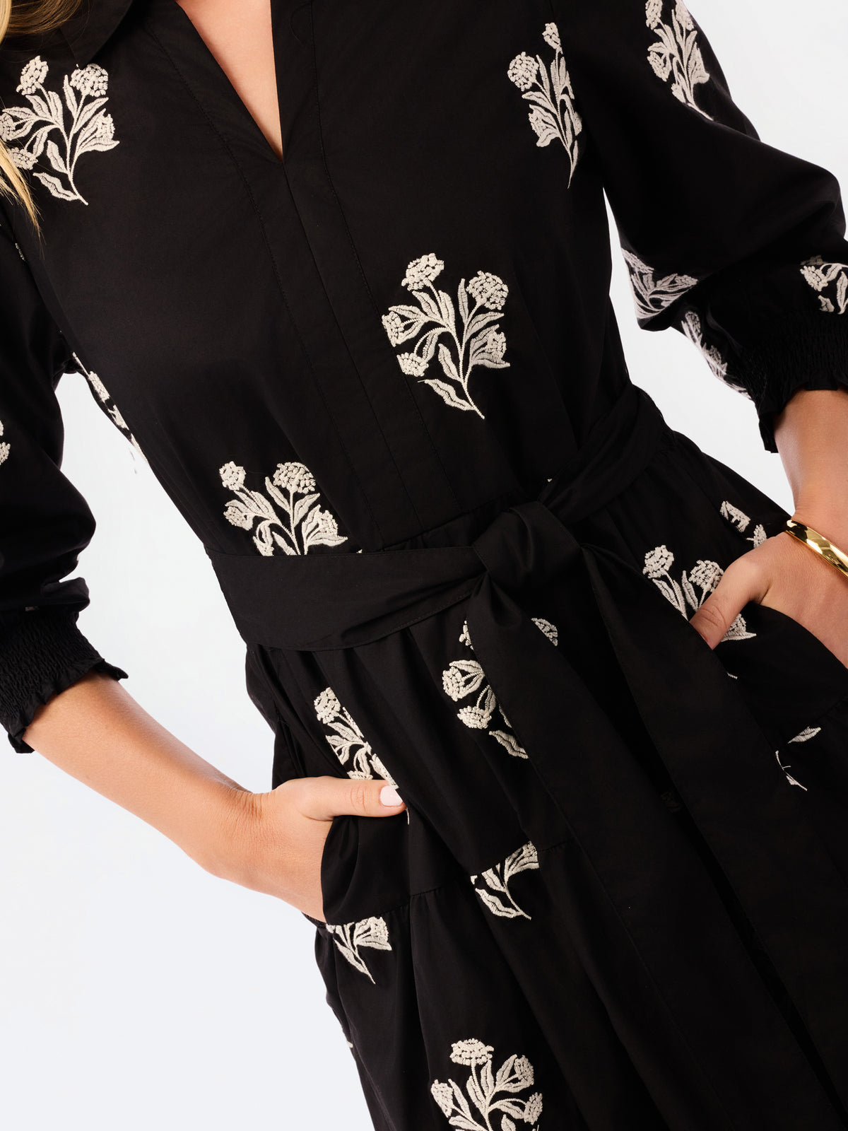 A close-up of a black long-sleeve dress with white floral embroidery, featuring a tie waist belt and side pockets, worn by a model.