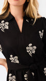 A close-up of a woman wearing a black long-sleeve collared dress with white floral embroidery and a tie waist, against a plain light background.