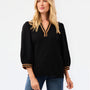 A blonde woman modeling a black long-sleeve blouse with gold trim on the V-neck and cuffs, paired with blue jeans, against a plain white studio background.