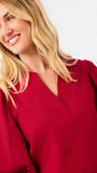 A woman wearing a red long-sleeve blouse with puff sleeves and a split neckline, paired with blue jeans, photographed against a plain white studio background.