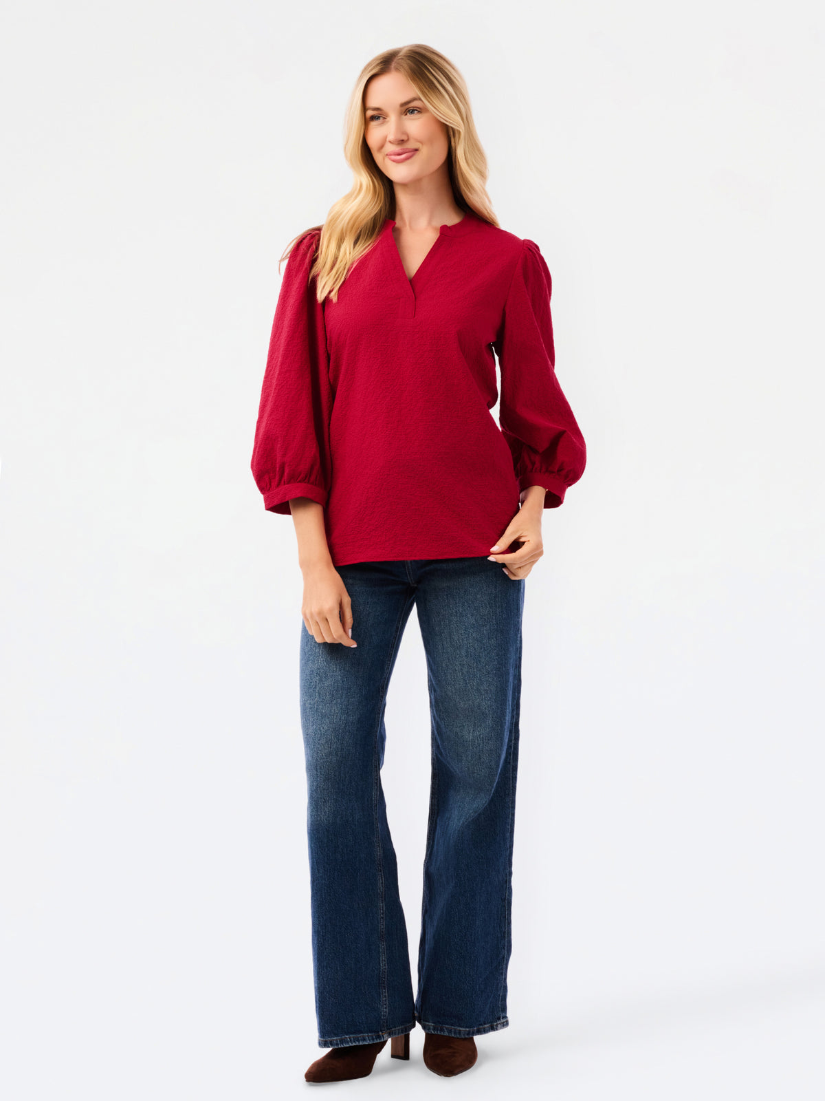 A woman wearing a red long-sleeve blouse with puff sleeves and a split neckline, paired with blue jeans, photographed against a plain white studio background.