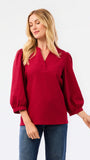 A woman wearing a red long-sleeve blouse with puff sleeves and a split neckline, paired with blue jeans, photographed against a plain white studio background.