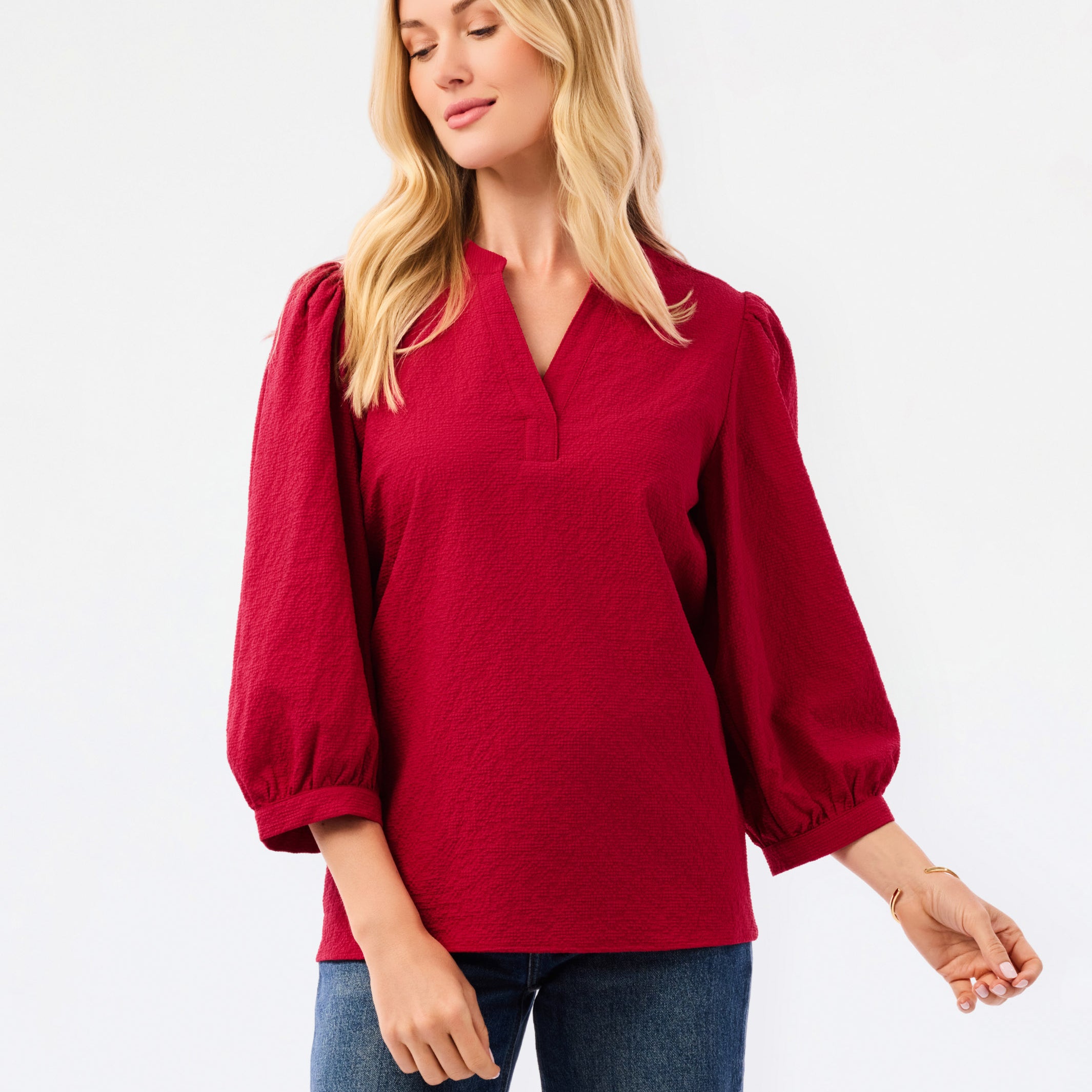 A woman wearing a red long-sleeve blouse with puff sleeves and a split neckline, paired with blue jeans, photographed against a plain white studio background.