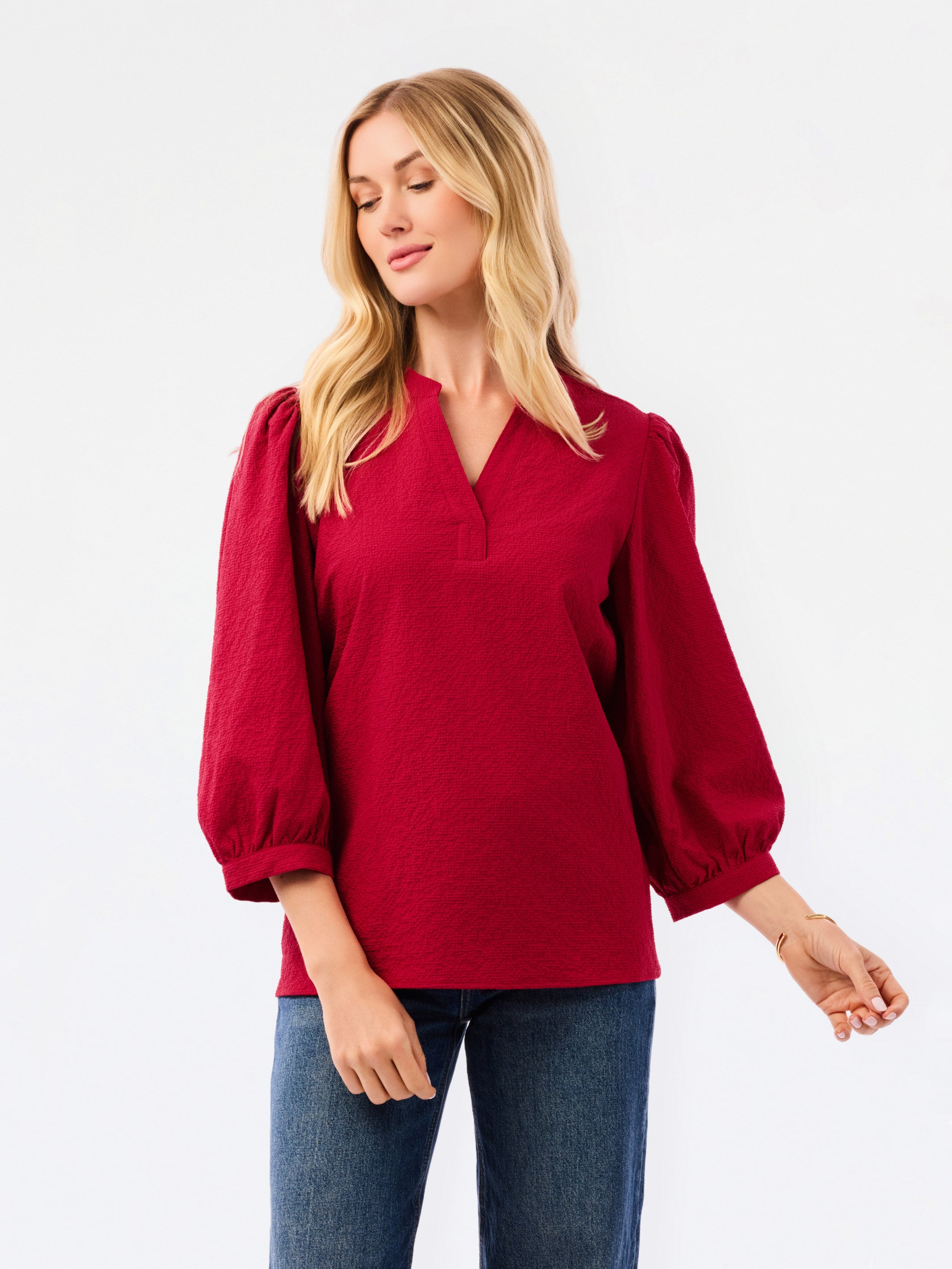 A woman wearing a red long-sleeve blouse with puff sleeves and a split neckline, paired with blue jeans, photographed against a plain white studio background.