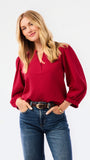 A woman wearing a red long-sleeve blouse with puff sleeves and a split neckline, paired with blue jeans, photographed against a plain white studio background.