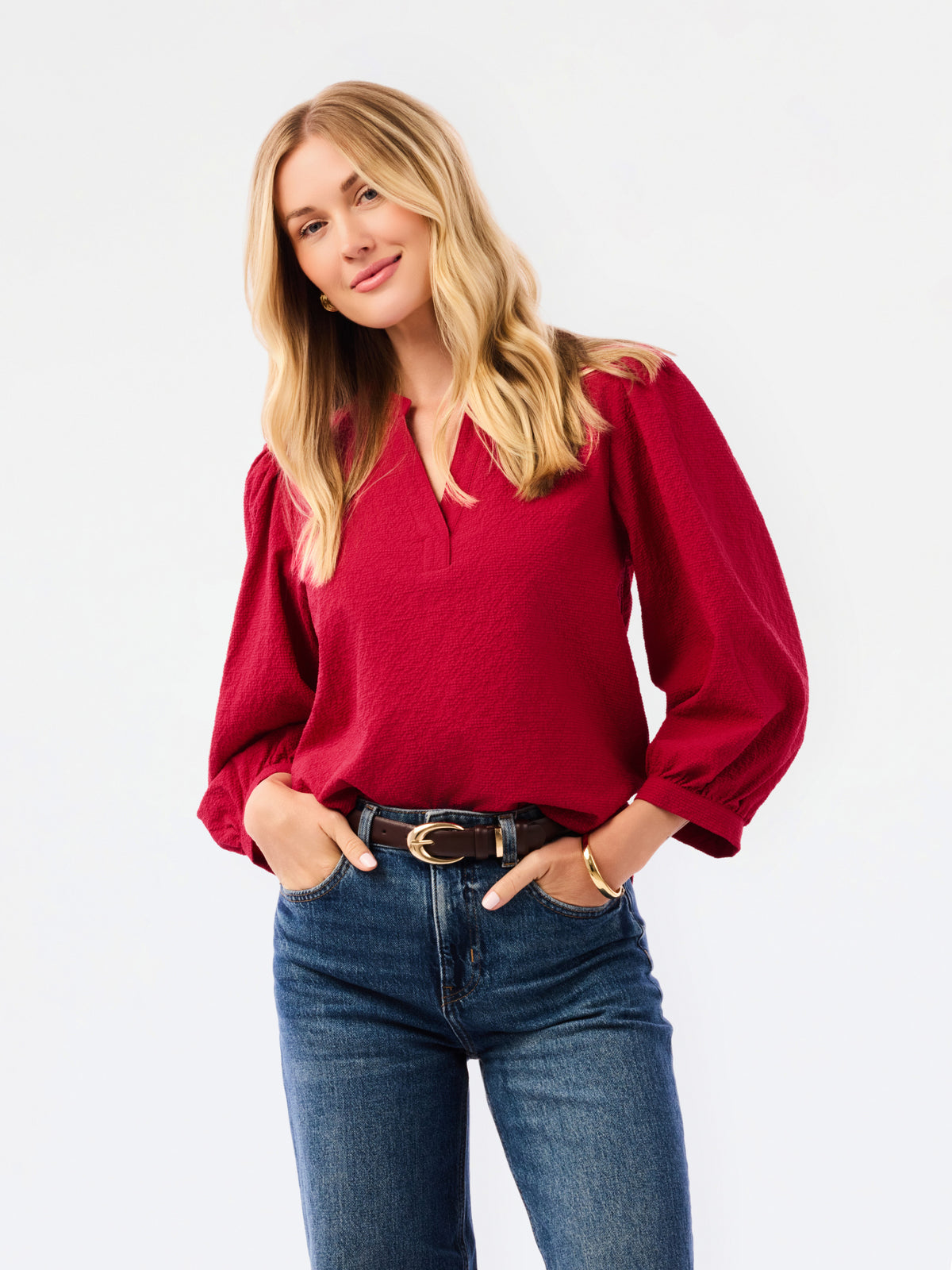 A woman wearing a red long-sleeve blouse with puff sleeves and a split neckline, paired with blue jeans, photographed against a plain white studio background.