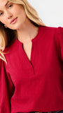 A woman wearing a red long-sleeve blouse with puff sleeves and a split neckline, paired with blue jeans, photographed against a plain white studio background.