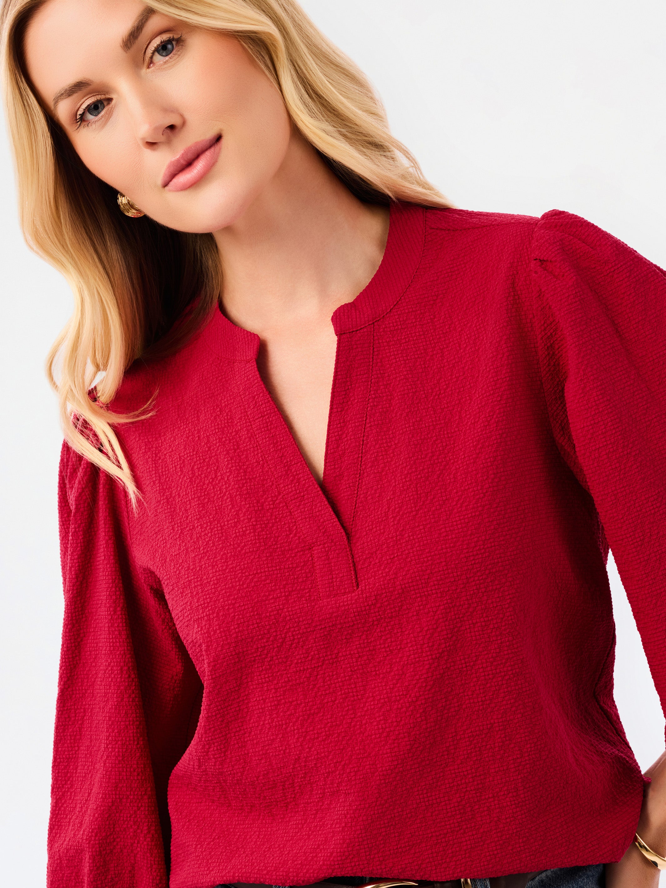 A woman wearing a red long-sleeve blouse with puff sleeves and a split neckline, paired with blue jeans, photographed against a plain white studio background.