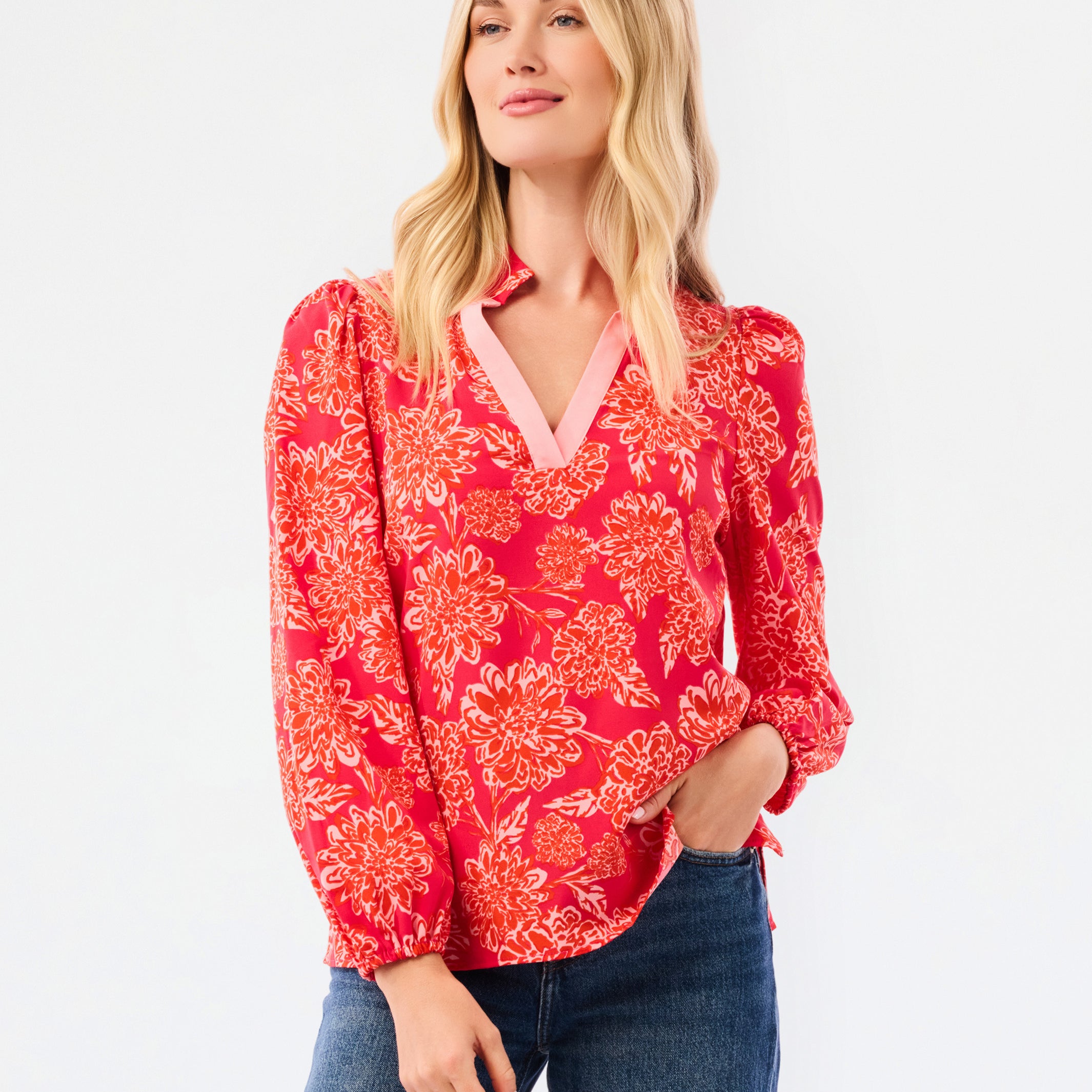 A woman modeling a red long-sleeve blouse with a white floral pattern and a pink inner V-neck, paired with blue jeans, against a plain white studio background.