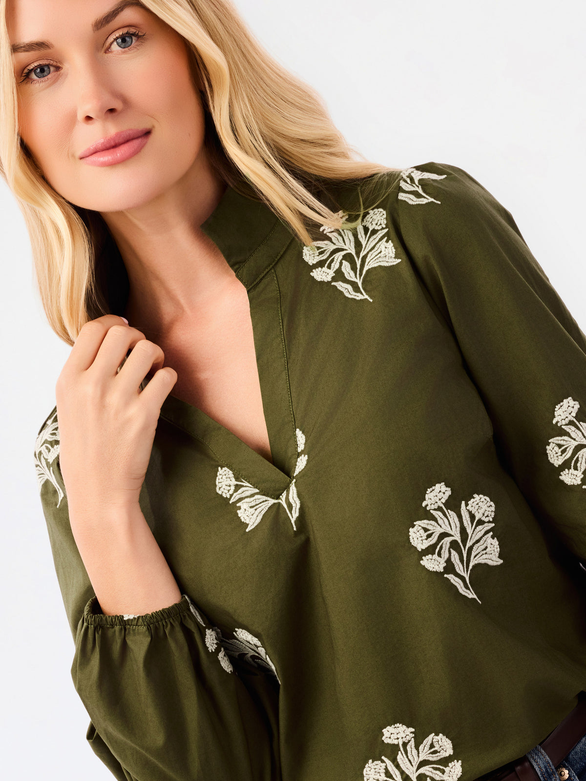 A close-up of a blonde woman wearing an olive green blouse with white floral embroidery, featuring a deep V-neck and long sleeves.