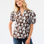 A woman modeling a brown short-sleeve blouse with a white floral print, paired with blue jeans, against a plain white studio background.