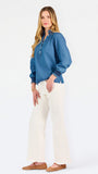 Blonde woman wearing a long sleeve denim shirt with navy blue trim and wearing creme pants