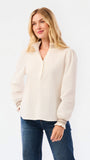 A blonde woman wearing a cream-colored long-sleeve blouse with lace trim at the collar and cuffs, paired with blue jeans, against a plain white studio background.