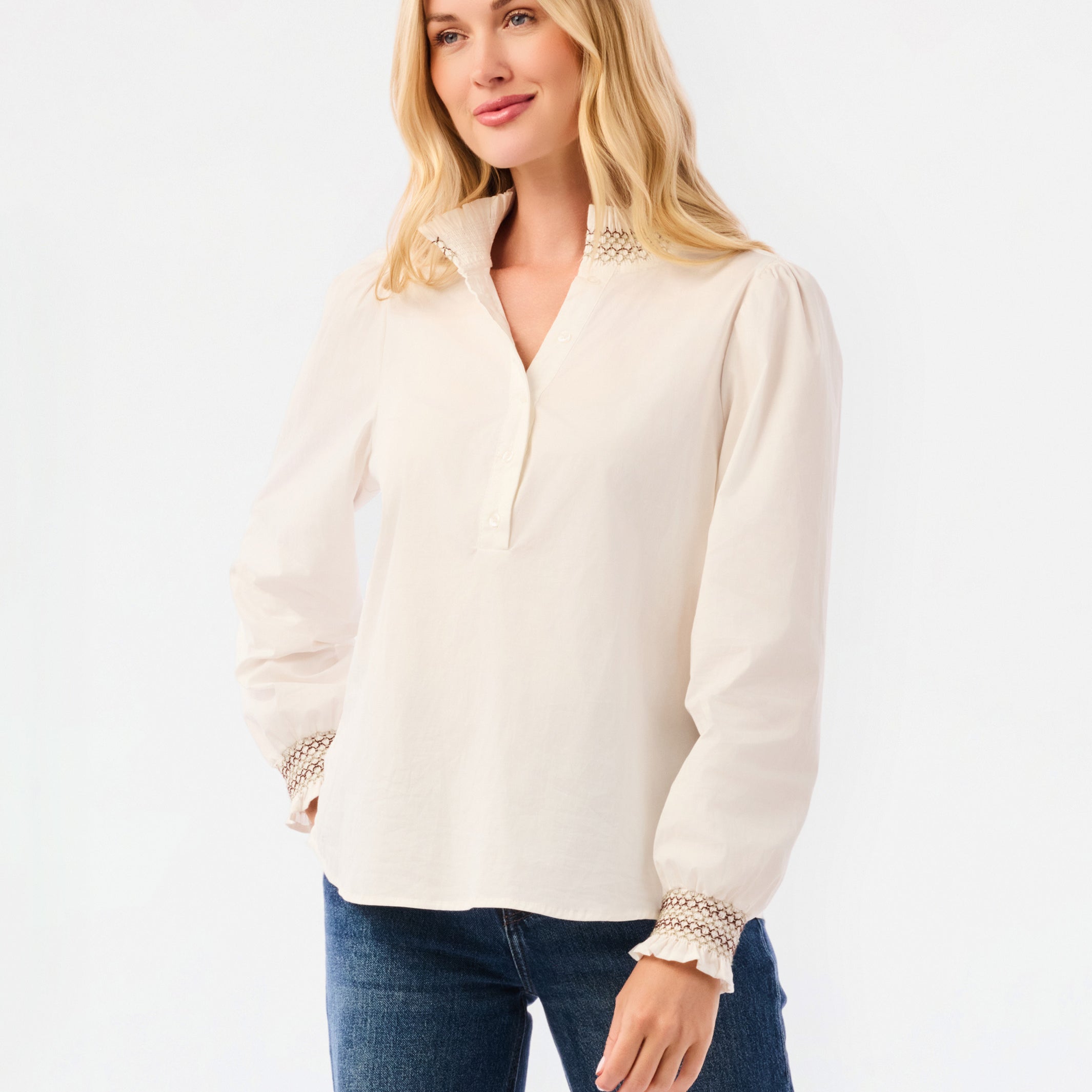 A blonde woman wearing a cream-colored long-sleeve blouse with lace trim at the collar and cuffs, paired with blue jeans, against a plain white studio background.