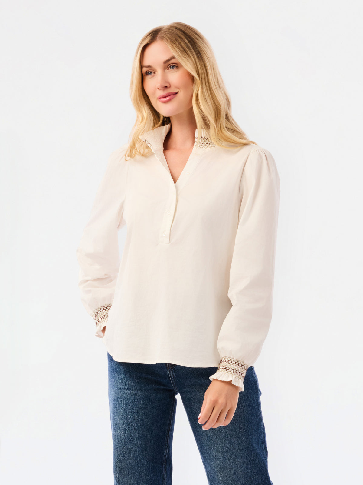 A blonde woman wearing a cream-colored long-sleeve blouse with lace trim at the collar and cuffs, paired with blue jeans, against a plain white studio background.