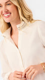 A blonde woman wearing a cream-colored long-sleeve blouse with lace trim at the collar and cuffs, paired with blue jeans, against a plain white studio background.