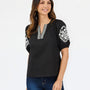 A woman modeling a black short-sleeve top with white floral embroidery on the sleeves and a white embroidered V-neckline trim, paired with blue jeans against a plain white background.