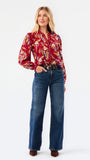 A woman modeling a red and beige abstract patterned top with blue jeans, photographed against a plain white studio background.