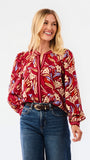 A woman modeling a red and beige abstract patterned top with blue jeans, photographed against a plain white studio background.