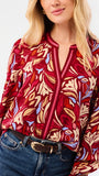 A woman modeling a red and beige abstract patterned top with blue jeans, photographed against a plain white studio background.