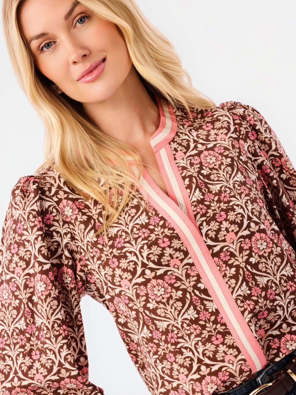 A smiling blonde woman models a long-sleeve patterned blouse with pink trim and blue jeans against a plain white studio background.