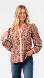 A smiling blonde woman models a long-sleeve patterned blouse with pink trim and blue jeans against a plain white studio background.