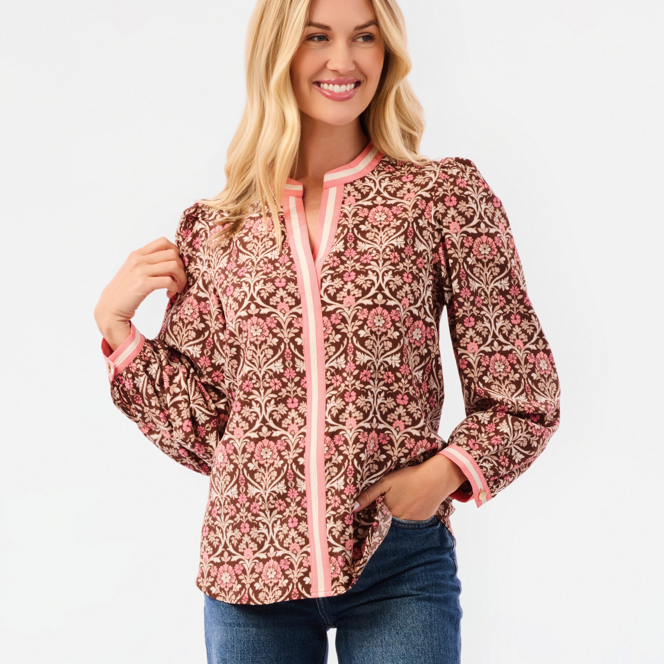A smiling blonde woman models a long-sleeve patterned blouse with pink trim and blue jeans against a plain white studio background.