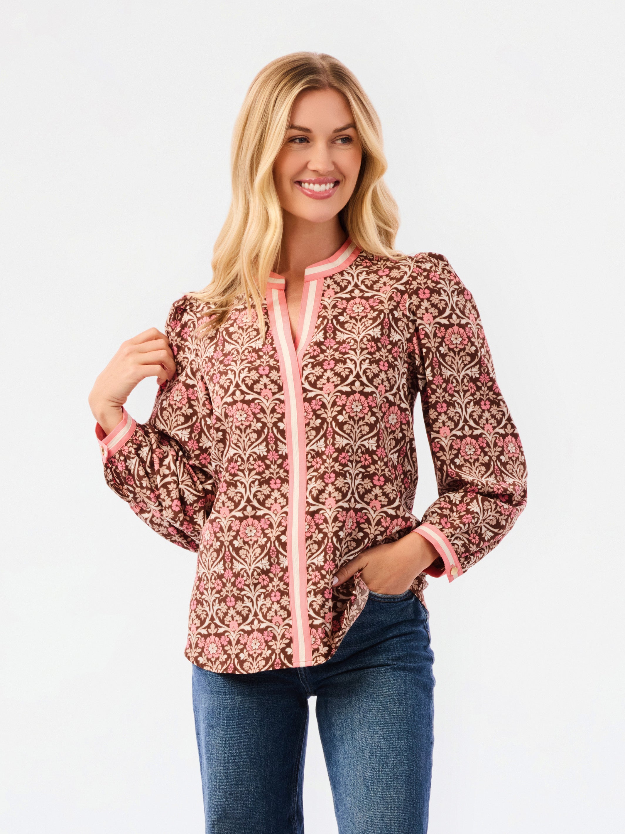 A smiling blonde woman models a long-sleeve patterned blouse with pink trim and blue jeans against a plain white studio background.
