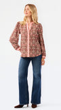 A smiling blonde woman models a long-sleeve patterned blouse with pink trim and blue jeans against a plain white studio background.