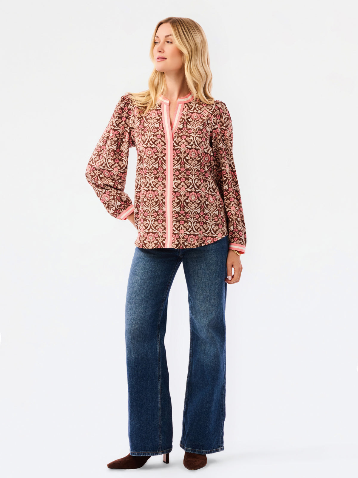 A smiling blonde woman models a long-sleeve patterned blouse with pink trim and blue jeans against a plain white studio background.